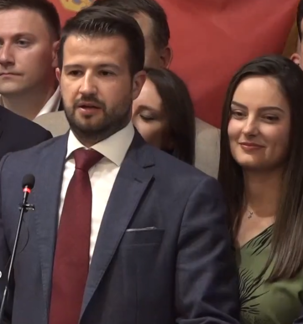 Ein Mann im Anzug und Krawatte steht an einem Rednerpult und hält eine Rede vor einer Menge mit einem Banner im Hintergrund, wahrscheinlich wegen eines Regierungsverbots von Social Media.