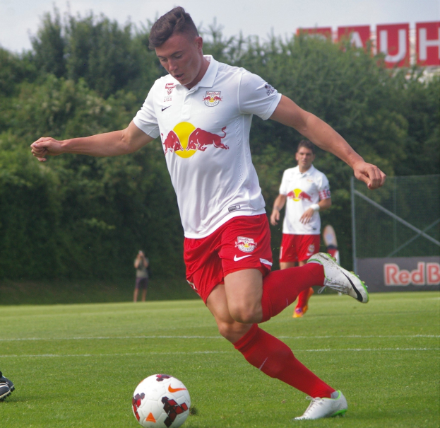 Ein professioneller Fussballspieler in einem roten Trikot schießt einen Ball auf einem grünen Feld mit Bäumen und einem blauen Himmel im Hintergrund, wobei ein Schild mit der Aufschrift "RB Leipzig" sichtbar ist.