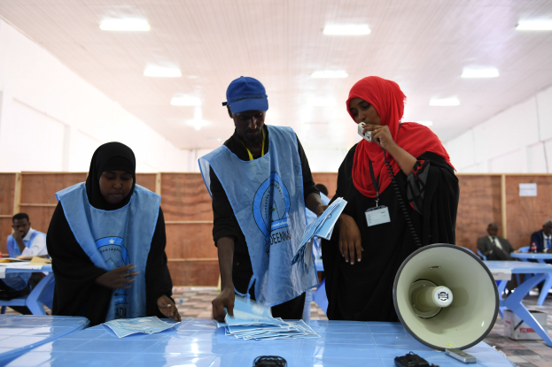 Eine Gruppe von Menschen steht um einen Tisch mit einem Megafon, Papieren und anderen Gegenständen herum, in einem Raum mit sitzenden und stehenden Personen und Deckenleuchten, was auf eine Wahllokal für die Stimmabgabe hinweist.