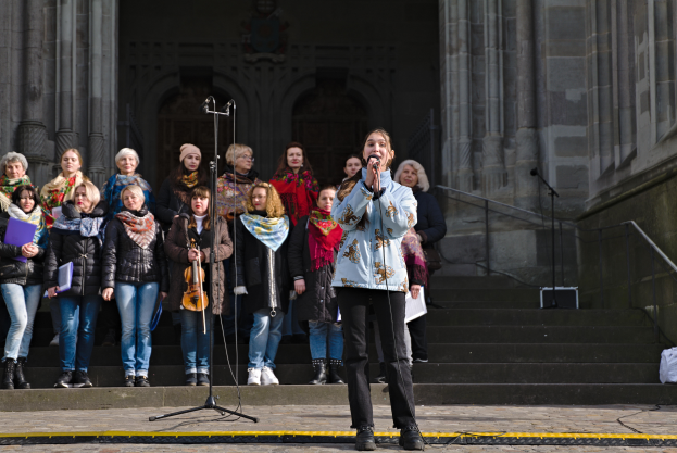 Eine Gruppe von Menschen, einige mit Musikinstrumenten, steht vor einem Gebäude mit Stufen und Geländern, während eine Frau in der Mitte in ein Mikrofon singt, das von Mikrofonstativen umgeben ist.