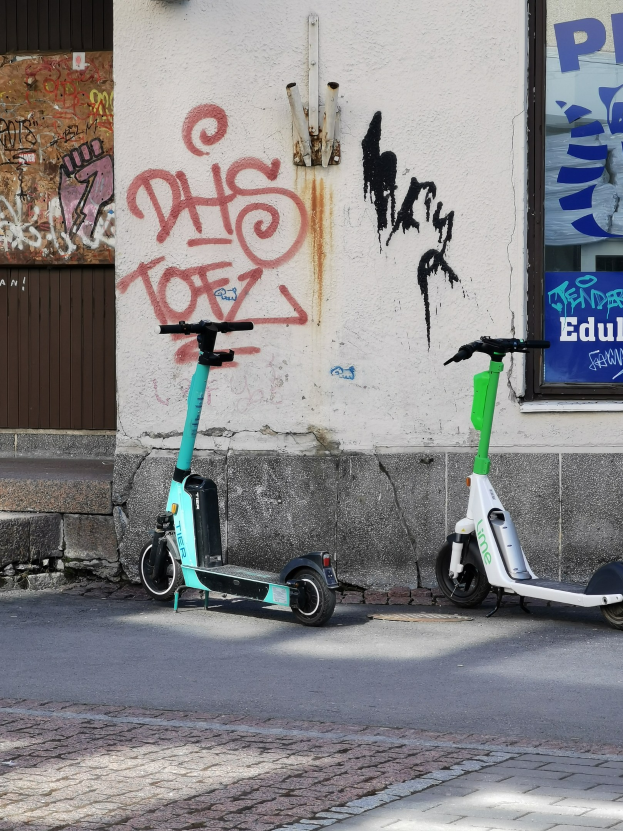 Zwei elektrische Roller nebeneinander auf einer Straße mit einer Graffiti-bedeckten Wand und einem Plakat im Hintergrund geparkt.
