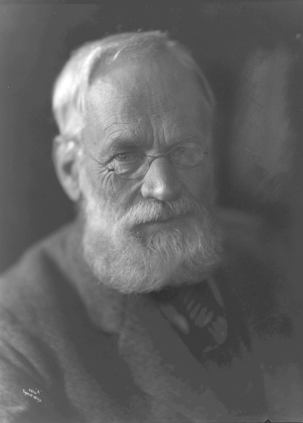 Alter Mann mit Vollbart und Brille, der dunkle Kleidung trägt und konzentriert in die Ferne schaut, vor einem dunklen Hintergrund.