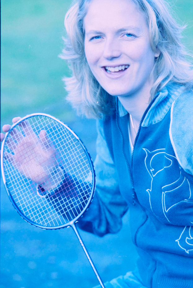 Eine Frau mit strahlendem Lächeln, die ein weißes Hemd und blaue Shorts trägt, hält einen Badminton-Schläger in der Hand und schaut konzentriert darauf, bereit zum Spiel.