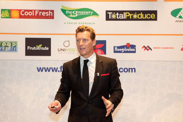 Ein Mann in einem schwarzen Blazer steht und hält ein Objekt, während er spricht, mit einem weißen Banner und einem weißen Board mit organisationalen Logos im Hintergrund.