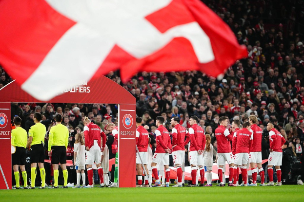 Eine Gruppe von Menschen auf einem Fußballfeld mit einer roten und weißen Flagge im Vordergrund, einem Bogen mit dem Text "Bayern Munich vs Bayern Munich Wett-Tipps & Vorschau" im Hintergrund und einer großen Menge im Stadion.