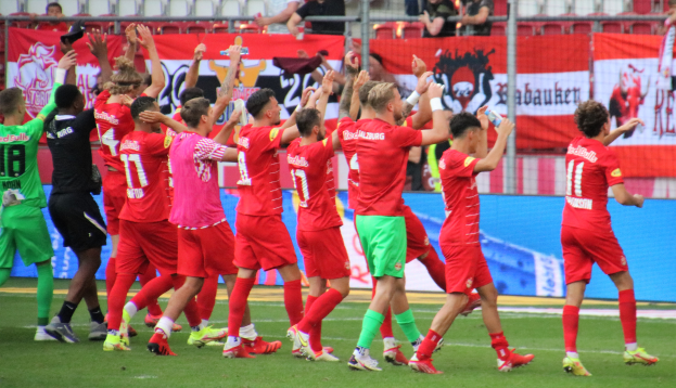 Eine Gruppe von Füßballspielern, die ein Tor feiern, einige halten Flaschen und andere klatschen, mit einem Zaun, Bannern, Metallständen, Stühlen und einer Decke mit Deckenlampen im Hintergrund.