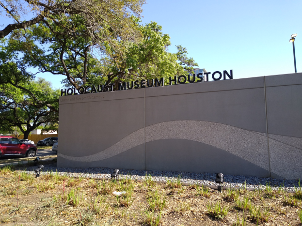 Außenansicht des Holocaust-Museums Houston mit eingravierter Textwand, Gedenksteine, Landschaftsgestaltung, Straßenverkehr, Straßenlaterne und bewölktem Himmel.