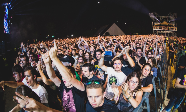 Menschenmenge auf einem Musikfestival mit erhobenen Händen, einige sitzen und andere stehen, umgeben von einem Metallzaun, mit Gebäuden und Lichtern im Hintergrund unter einem dunklen Himmel.