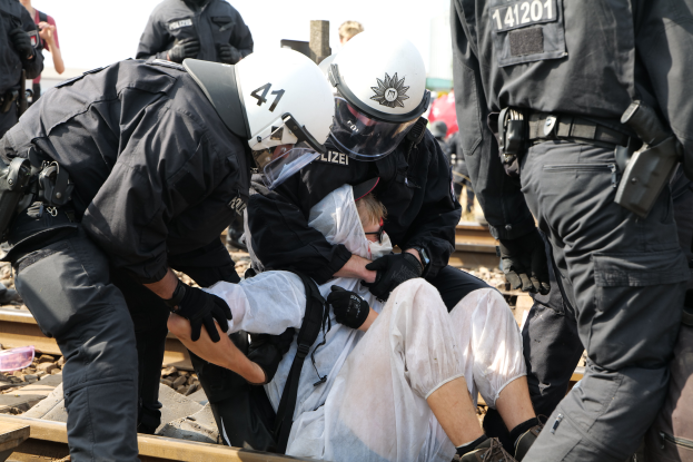 Eine Gruppe von Polizisten in schwarzer Einsatzkleidung verhaftet einen Mann in einem weißen Kleid auf den Bahnschienen, mit anderen Menschen und dem Himmel im Hintergrund.