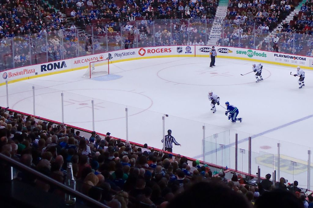 Vier Männer beim Eishockey auf einem Eisstadion, mit zwei in der Nähe des Tors und Zauns stehend, Zuschauern auf Stühlen im Vorder- und Hintergrund sowie einer Treppe im Hintergrund.