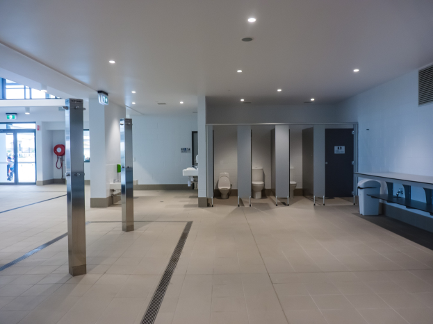Große ßeffentliche Toilette mit mehreren Toiletten, Säulen, einem Mülleimer, einer Tür, einem Schild, einem Feuerlöscher und Deckenleuchten.