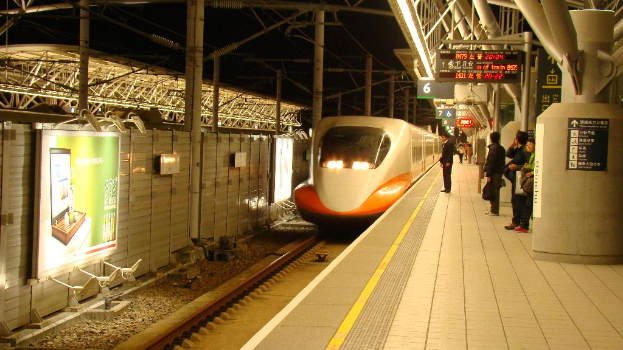 Eine Gruppe von Menschen steht auf einem Bahnsteig mit einem Zug auf den Schienen, eine Wand mit einer Tafel links und Pfosten mit Tafeln rechts.