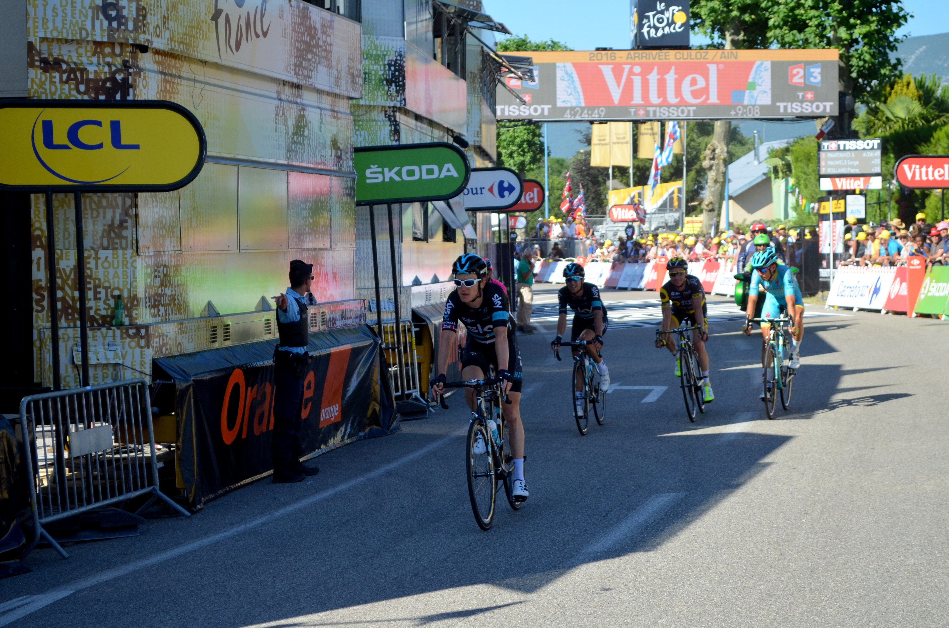 Eine Gruppe von Radfahrern in Helmen und Schutzbrillen fährt eine Straße mit Barrieren, Schildern und Tafeln mit Text hinunter, während eine Person auf der linken Seite steht; Bäume, Gebäude, Flaggen und Pfosten säumen die Strecke unter einem klaren blauen Himmel.