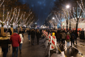 Eine Menschenmenge, die nachts eine Straße entlanggeht, mit Bäumen und Gebäuden, die von festlichen Lichtern beleuchtet werden, unter einem bewölkten Himmel.