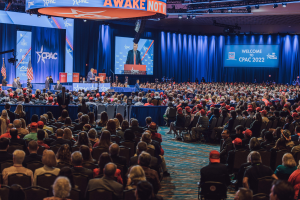 Eine große Menschenmenge sitzt auf Stühlen, die einer Bühne zugewandt sind, mit ein paar Menschen darauf, blaue Vorhänge und ein Bildschirm im Hintergrund und eine orangefarbene Tafel oben, die das CPAC 2020-Event am 25. Oktober anzeigt.