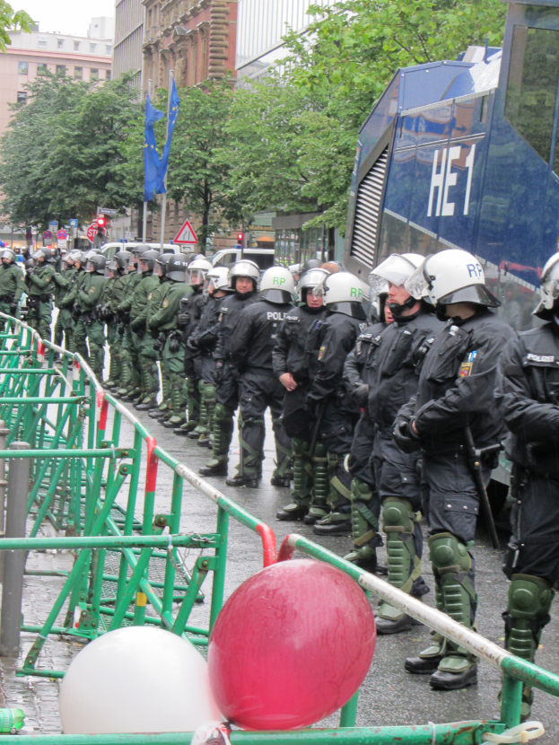 Eine Gruppe von Polizisten in Helmen und mit Waffen vor einer Absperrung auf einer Straße mit Geländern und Ballons, mit Bäumen, Gebäuden, Flaggen und einem klaren blauen Himmel im Hintergrund.