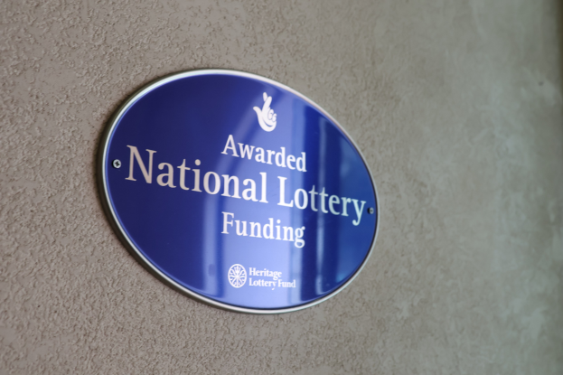 Blauer rechteckiger Schild an einer Gebäudewand mit weißem Hintergrund und schwarzer fetter Schrift, die "Mit Nationaler Lotterie-Förderung ausgezeichnet" lautet.
