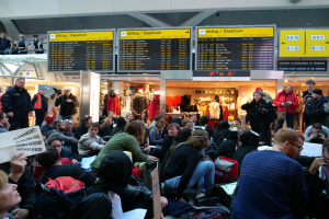 Eine große Gruppe von Menschen in einem Flughafen, einige mit Taschen und Papieren sitzend, andere stehend, mit Texttafeln, Schaufensterpuppen in Kleidern und Deckenlichter im Hintergrund, was auf eine Protestaktion hindeutet.