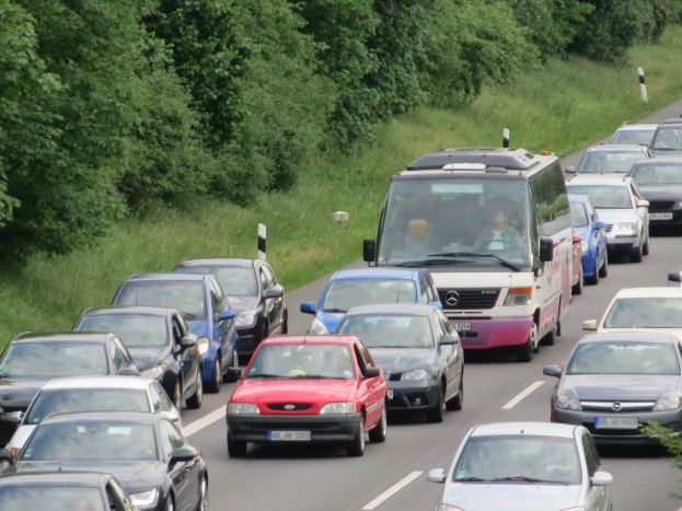 Ein Stau auf einer Autobahn mit mehreren Autos und einem Lieferwagen, Menschen sind in den Fahrzeugen zu sehen und im Hintergrund gibt es Bäume und Gras.