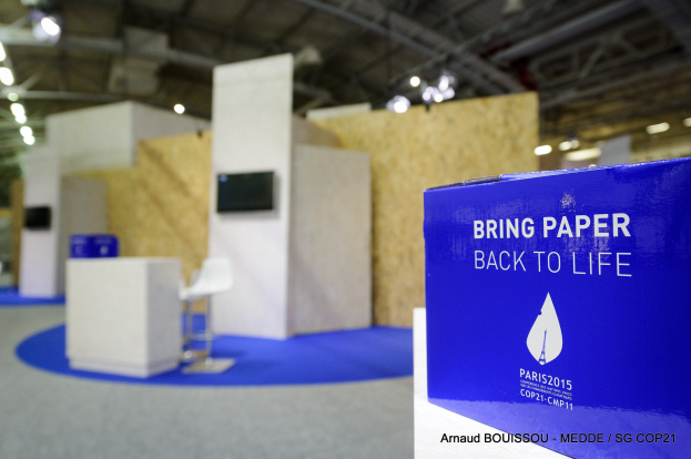 Messestände mit einem blauen und weißen "Bring Paper Back to Life"-Schild auf der rechten Seite, das einen blauen und weißen Hintergrund mit Stühlen, Tischen, einer Wandfernseher und Deckenlampen zeigt.