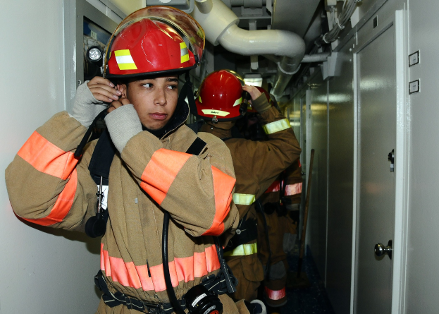 Gruppe von Feuerwehrleuten in Uniform, die zusammen in einem Trainingsraum mit Rohren und Equipment im Hintergrund stehen.