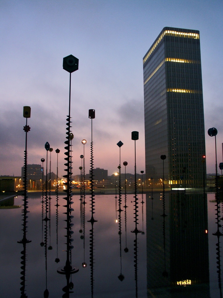 Mehrere Masten und Gebäude mit Lichtern unter einem bewölkten Himmel, mit ihren Spiegelungen.