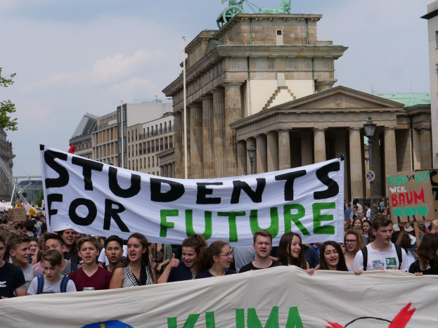 Gruppe von Schülern marschiert in Berlin mit einer bunt bemalten "Students for Future"-Schriftzug vor Gebäuden, Bäumen und Himmel.