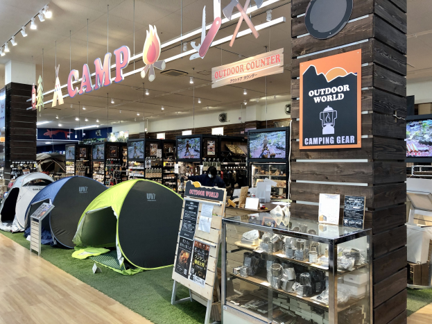 Ein Ladeneinrichtungsbild mit verschiedenen Campingausrütstücken, darunter Zelte, Regalen, einer Glasvitrine mit Gegenständen, Fernsehern, Deckenleuchten und Informationsschildern.