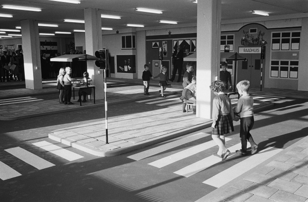 Schwarzes und weißes Foto von Kindern, die eine Straße an einer Ampel überqueren, mit Menschen auf Bänken, einem Verkehrssignalmast im Vordergrund und Säulen mit Deckenleuchten im Hintergrund.