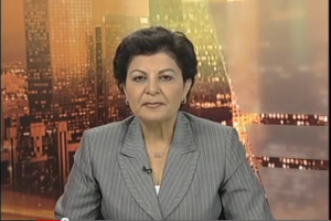 Eine Frau in einem Business-Outfit sitzt vor einem Fernseher mit einem News-Ticker unten, mit ein paar Gebäuden im Hintergrund.