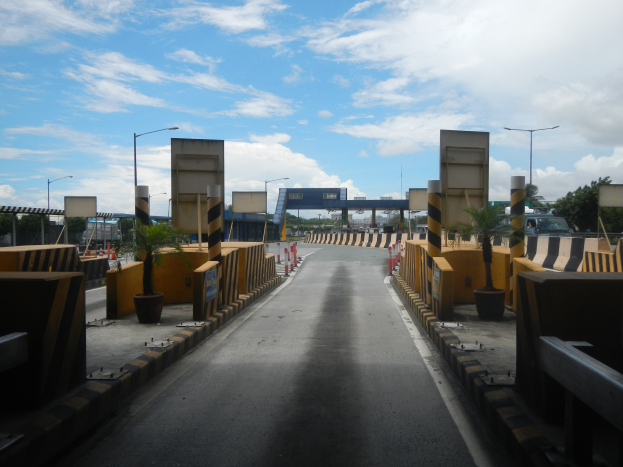 Straße mit einer Mäßstation, Absperrpfählen, Verkehrskegeln, Topfpflanzen, Schilder, Straßenmasten, Bäume und eine bewölkte Himmel.