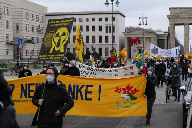 Eine große Gruppe von Menschen marschiert auf einer Straße, hält Schilder und Fahnen und demonstriert gegen Atomkraft in Deutschland, mit Fahrzeugen, Gebäuden, Laternenpfählen, Bäumen und einem Tor im Hintergrund.