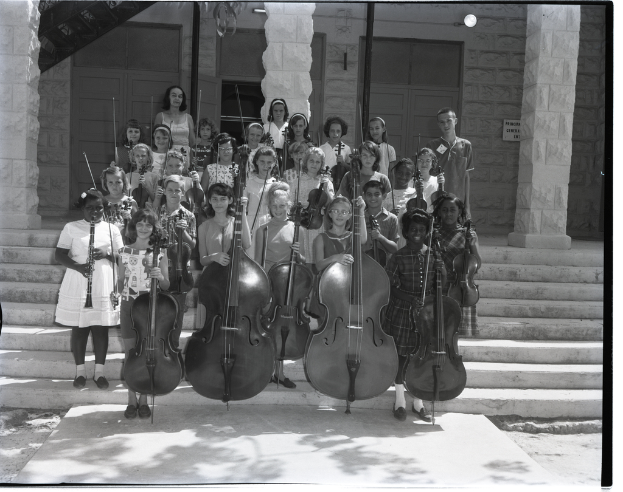 Eine Gruppe von Kindern steht auf einer Treppe und hält verschiedene Musikinstrumente wie Celli, Geigen und Trompeten, mit Säulen und einer Wand mit Türen im Hintergrund.