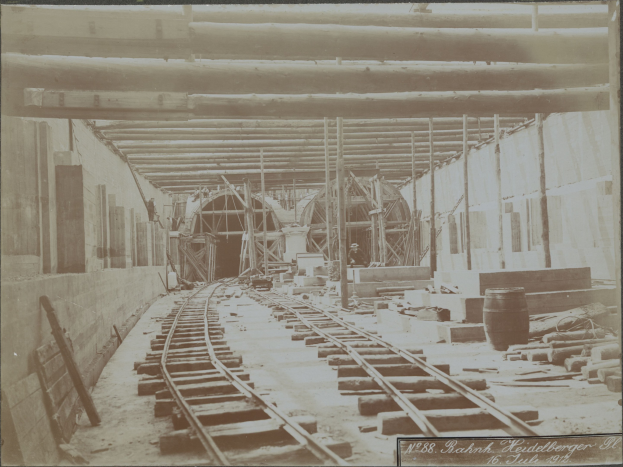 Schwarz-weiß-Foto einer Bahngleise im Inneren eines Gebäudes unter construction, mit verstreuten Holzobjekten, Fässern, Wänden, Pfählen und Text am unteren Rand.