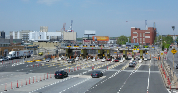 Eine belebte Straße mit verschiedenen Fahrzeugen, darunter Autos, Lkw und ein Landfahrzeug, umgeben von Gebäuden, Straßenlaternen, Bäumen und anderen urbanen Elementen unter einem bewölkten Himmel.