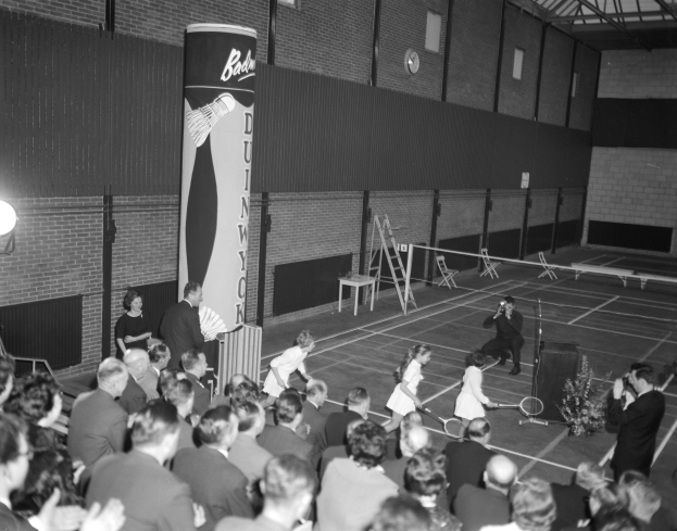 Ein Schwarz-Weiß-Foto einer Gruppe von Menschen, die Tennis in einer Turnhalle spielen, wobei einige stehen und andere auf Stühlen sitzen und Schläger halten, und eine Uhr an der Wand im Hintergrund.