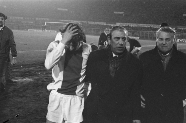 Eine Gruppe von Männern steht zusammen auf einem Fußballfeld, sieht frustriert aus und hat ein Stadion im Hintergrund, dargestellt in Schwarz-Weiß.