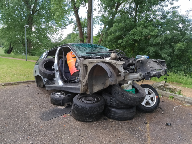 Ein umgestürzter Wagen am Stra├čenrand mit verstreuter Trümmern, umgeben von Gras, Pflanzen, Büschen, einer Stra├čenlaterne und einem bew├Âlktem Himmel.