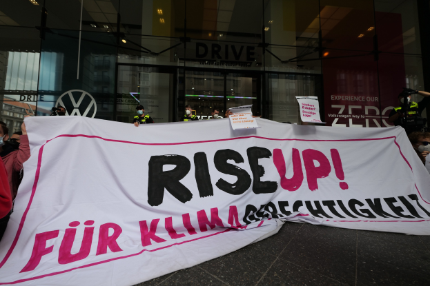 Eine Gruppe von Menschen in Masken, die eine "Steh Auf"-Plakette und Schilder vor einem Glasgebäude bei Nacht halten.