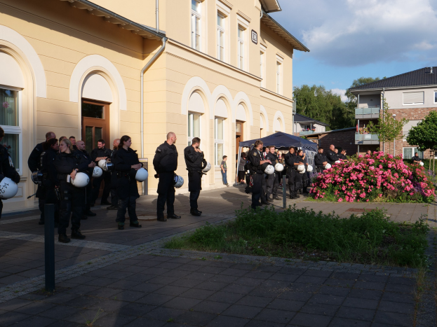 Gruppe von Polizisten vor einem Gebäude mit Fenstern und Türen stehend, Helme haltend, in der Nähe eines Zeltes, umgeben von Pflanzen, Blumen, Gras, einem Straßenpfahl, Bäumen und einem bewölkten Himmel.