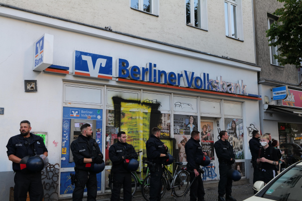 Polizeibeamte in Uniform stehen vor einer Berliner Volksbank-Filiale, einige halten Helme in den Händen, rechts ein Auto und links ein Baum; im Hintergrund sind Gebäude mit Fenstern und textbedruckten Schildern zu sehen.