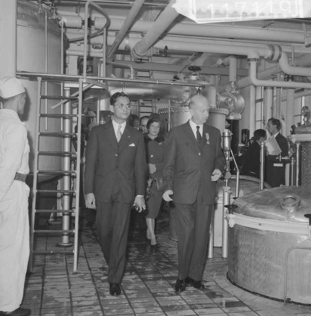 Eine Gruppe von Männern in Anzügen und Krawatten geht durch eine schwarz-weiße Fabrik, mit einer Leiter auf der linken Seite und Industriegeräten im Hintergrund.