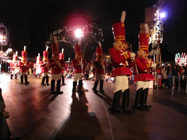 Menschen in roter und weißer Kleidung stehen mit Musikinstrumenten in den Händen. Im Hintergrund sitzen weitere Menschen auf dem Boden. Rechts sind Absperrpoller und eine Werbetafel mit Lichtern sowie ein geschmückter Baum und ein Gebäude zu sehen. Links sind beleuchtete Spiele zu erkennen, darüber ein dunkler Himmel und darunter ein Gehweg.