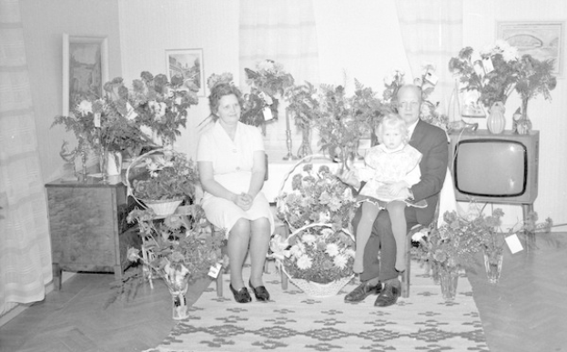 Schwarz-weiß-Foto einer Familie aus drei Personen - einem Mann, einer Frau und einem Kind - auf einem Sofa sitzend mit Blumenvasen auf benachbarten Tischen und einem Teppich auf dem Boden.