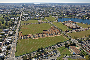 Luftaufnahme einer Stadt mit Gebäuden, Bäumen, Straßen, Fahrzeugen, Wasser und Wolken, die einen großen zentralen Platz zeigt, der als Parkgebiet vorgeschlagen ist.