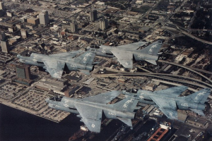 Vier Kampfflugzeuge fliegen über einer Stadt mit sichtbaren Gebäuden, Straßen, Fahrzeugen und Wasser, mit Text am unteren Bildrand.