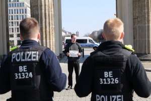 Drei Polizeibeamte stehen vor einem Gebäude, ein Mann in der Mitte hält ein Papier, flankiert von Säulen, während Fahrzeuge, Menschen, Bäume und Gebäude den Hintergrund unter einem klaren Himmel ausfüllen.