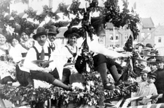 Eine Gruppe von Menschen in traditioneller bayerischer Tracht fährt auf einem mit Laub geschmückten Festwagen in einem Umzug, einige halten Musikinstrumente, mit Gebäuden und einem klaren Himmel im Hintergrund, dargestellt in Schwarz-Weiß.
