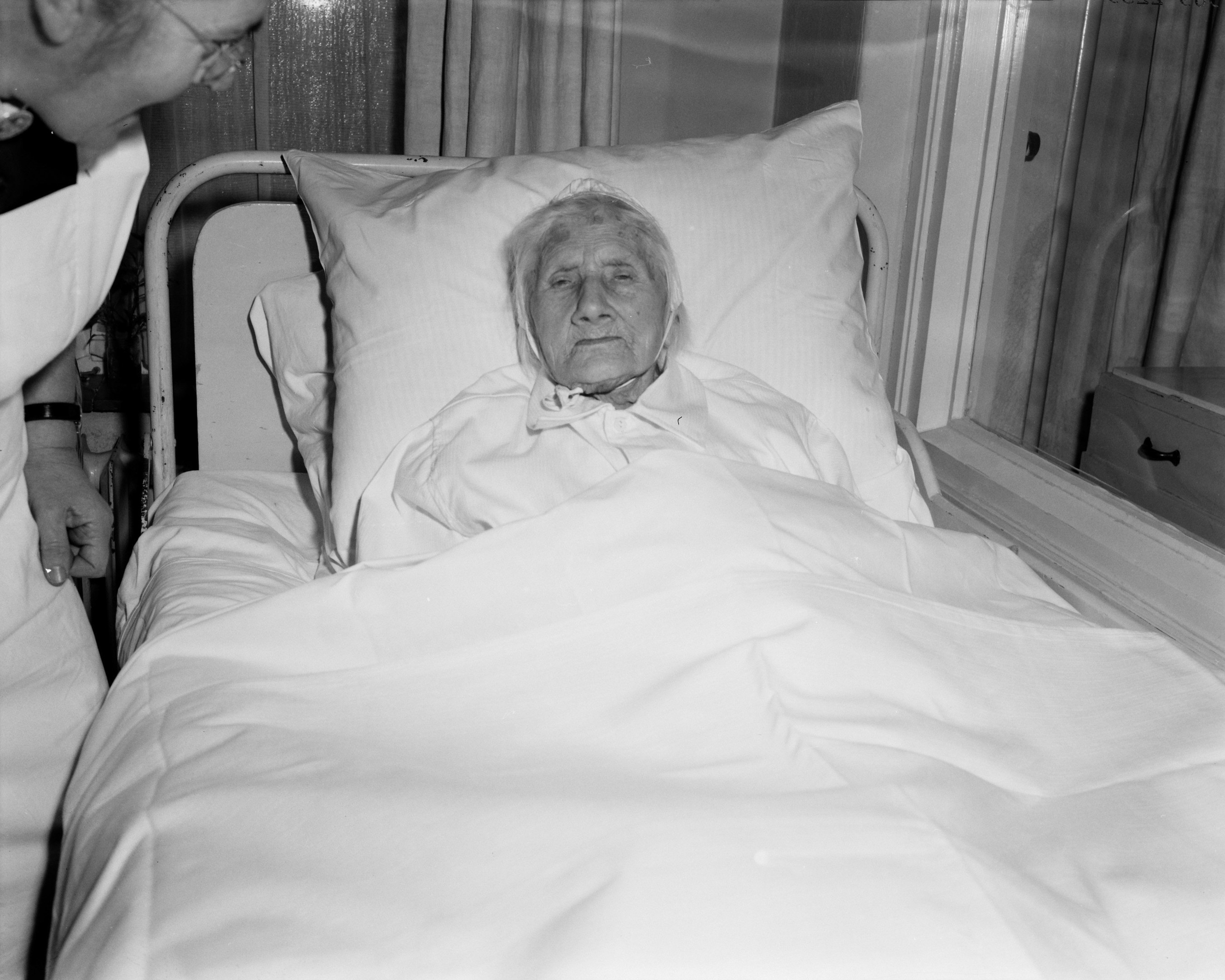 Ein älteres Weib liegt in einem Krankenhausbett mit einer weißen Decke und einem Kissen, während eine Krankenschwester daneben steht, mit einem Fenster mit Vorhängen im Hintergrund.