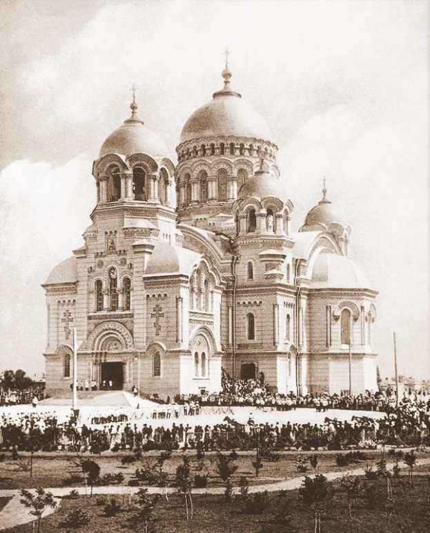 Schwarz-weiß-Fotografie der Erlöserkirche am Blutenstrom in St. Petersburg, Russland, mit Menschen, die sich um das prunkvolle Gebäude versammeln, Bäume und Pfosten im Vordergrund und einem klaren Himmel im Hintergrund.